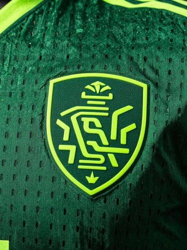 Macro close-up of a football jersey fabric, showcasing intricate ventilation mesh patterns and a heat-applied silicone crest under dramatic stadium lighting, rich green and neon accents