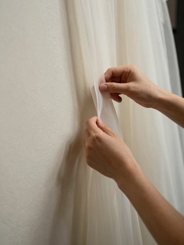Close-up of a fashion creator's hands arranging sheer fabric against a pale plaster wall, north-facing daylight, fine textile texture visible, compositional negative space to the left