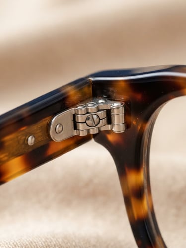 Macro close-up of premium tortoiseshell optical frame hinges, warm champagne background, sharp focus on craftsmanship, bright studio lighting