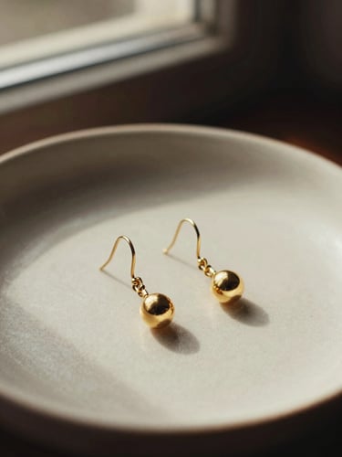 An elegant pair of gold-plated drop earrings resting on a textured ceramic dish, warm shadows.