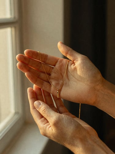 Close-up of hands holding a delicate gold necklace in warm golden-hour window light, soft textured linen background, shallow depth of field, 35mm film