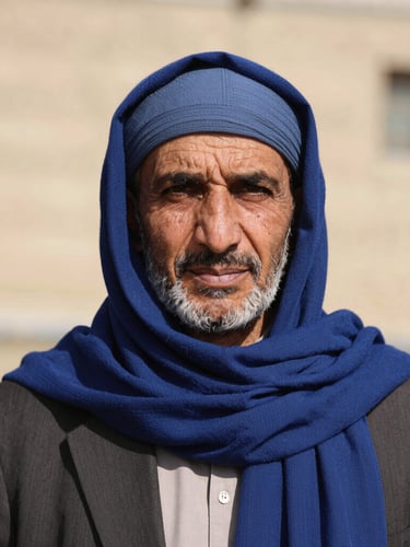 Dignified medium-close portrait of an Afghan community leader in natural bright daylight, direct eye contact, wearing a deep blue scarf, warm neutral background, sharp focus, 35mm