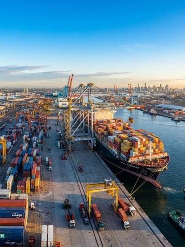 Wide panoramic shot of a cargo ship being loaded with containers at a bustling port at sunrise, global trade routes faintly overlaid, bright natural daylight, clear sky.