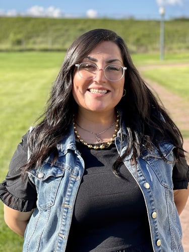 Smiling woman with dark hair and glasses wearing a denim vest. Cristina Adame