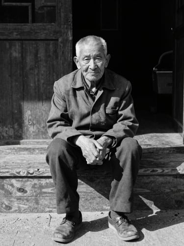 A high-contrast monochrome environmental portrait of an elderly resident sitting on a wooden stoop in harsh midday sun, deep shadows, gritty 35mm film grain.