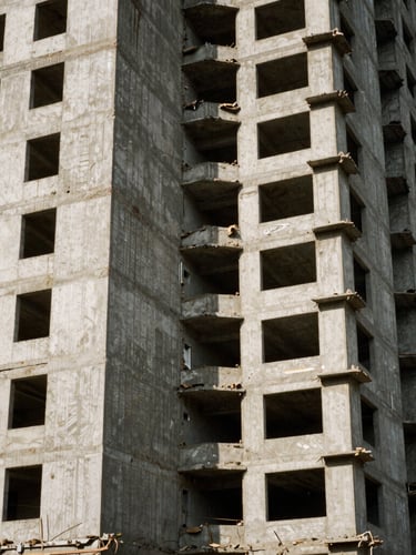 Gritty medium-format documentary photograph of a half-demolished concrete high-rise, harsh daylight casting sharp shadows, visible film grain, muted earth tones.
