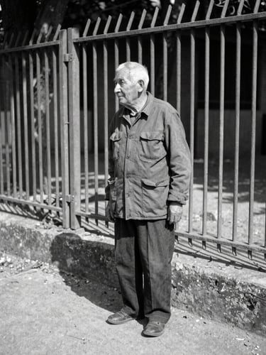 Documentary photo of an elderly resident standing near a weathered steel fence, harsh midday sun, sharp shadows on concrete, 35mm grain.