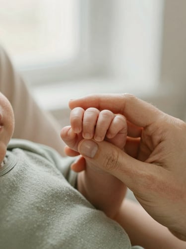 Close-up detail of a mother gently holding a newborn baby's tiny hand, soft natural window light, warm beige and sage green tones, shallow depth of field