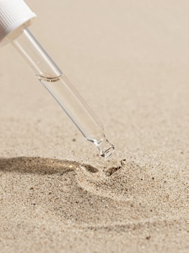 Macro photograph of soft warm sand texture with gentle shadows, light-colored clinical glass dropper reflecting soft daylight, minimalist and clean