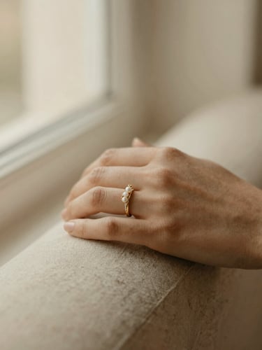 Close-up of a woman's hand wearing an elegant gold-plated bridal ring with subtle pearl accents, resting on soft beige linen, warm window light, shallow depth of field, muted cream and champagne tones, 35mm
