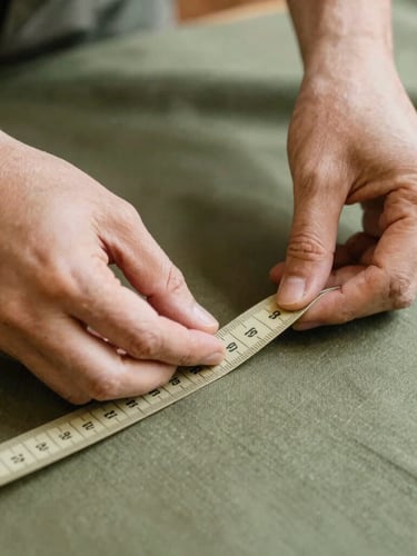 Close-up of artisan hands carefully measuring a soft olive green fabric with a classic tailor tape, warm natural light, shallow depth of field, detailed texture of the textile.