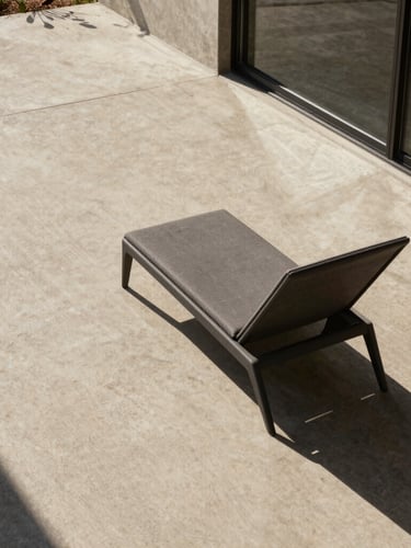 Wide architectural environmental shot, hard direct sunlight casting dramatic shadows on a raw concrete deck, a single charcoal structural lounge chair positioned near a glass threshold, medium format.