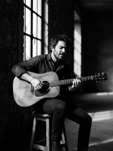 Moody monochrome portrait of Guillem MiTchel with his acoustic guitar in a quiet, architectural space with dramatic natural window light and deep shadows.