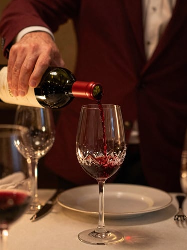 Close-up of a sommelier pouring red wine into a crystal glass at a finely set table, warm low-key ambient lighting, rich maroon and gold accents, shallow depth of field, 35mm