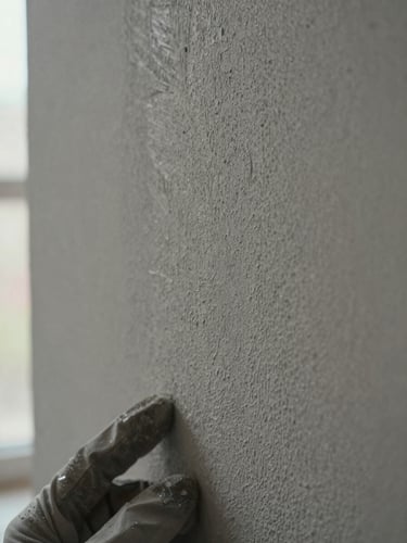 Extreme close-up of a plastered wall surface under diffuse north-facing window light, fine trowel marks visible in the matte greige finish, a gloved hand resting at the lower left edge, shallow depth of field emphasising the texture grain