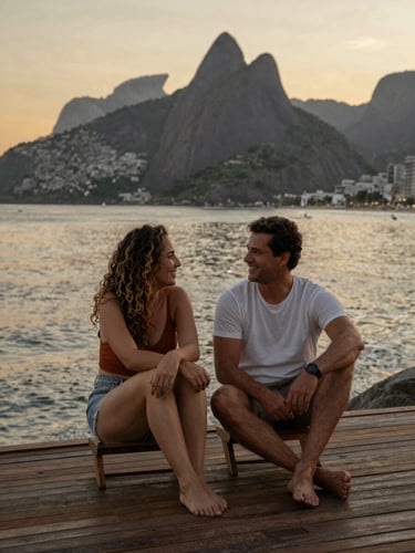 Fotografia profissional dos fundadores Fernanda Camargo e Wagner Rodrigues em Angra dos Reis, sorrindo em um deck de madeira ao entardecer, com o mar calmo e as montanhas ao fundo, luz dourada suave, enquadramento editorial de meio corpo.