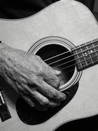 Close-up of weathered hands pressing down on acoustic guitar strings, gritty black and white photography, high-contrast chiaroscuro lighting, deep shadows, raw texture