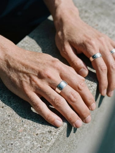 Close-up of hands with silver rings resting on a raw concrete ledge, high-contrast shadows, grain of medium-format film.