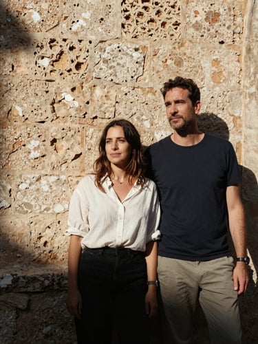Cinematic medium-format portrait of a couple standing by an ancient stone wall in Mallorca, harsh midday sun casting deep shadows, warm Mediterranean light, raw candid emotion, look away from camera.