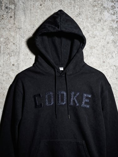 Tight crop gritty street photography of heavy embroidery on a black hoodie, harsh direct flash, high-contrast shadows, raw concrete texture background