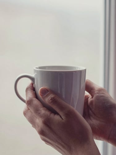 Close-up of hands holding a warm ceramic mug near a window, soft diffused morning light, muted pastel tones, shallow depth of field, 35mm