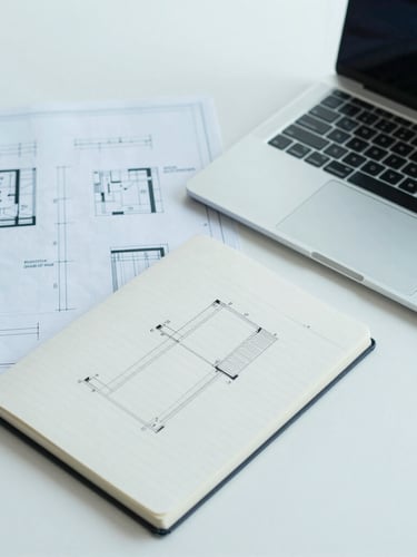 Overhead desk shot of a clean minimalist workspace. Technical blueprints, a sleek laptop, a notebook with structured architectural diagrams. Cool blue and teal lighting, sharp focus, professional editorial photography.