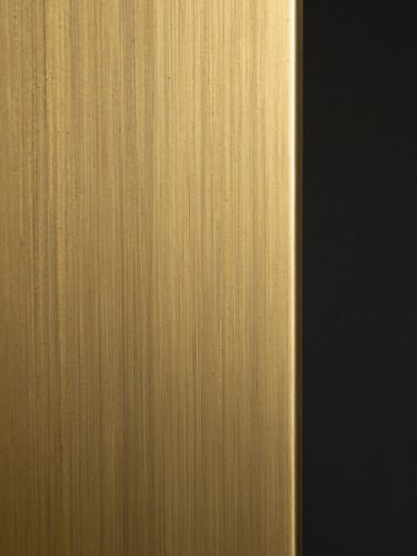 Extreme close-up of hand-finished brass panel surface under tungsten studio light, showing grain direction and subtle patina transition across the metal, tight crop isolating surface texture, dark background