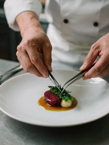 Editorial close-up of a chef's hands carefully plating a delicate dish with tweezers in a professional kitchen, moody natural daylight, shallow depth of field, 35mm film.