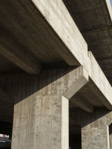 Vista de ángulo bajo de una estructura de hormigón visto con líneas geométricas perfectas, luz solar rasante que resalta la textura rugosa del encofrado y sombras marcadas.