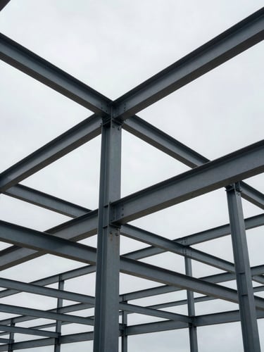 Wide shot from below looking up at a completed industrial steel frame structure, heavy I-beams and bolted connections visible in sharp detail, overcast natural daylight, no workers, no people, scale conveyed by the geometry of the frame against a grey sky