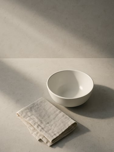 Close-up overhead composition of a minimalist kitchen counter — a single white ceramic bowl, a folded linen cloth, and cool morning light raking across pale stone surface, vast negative space above