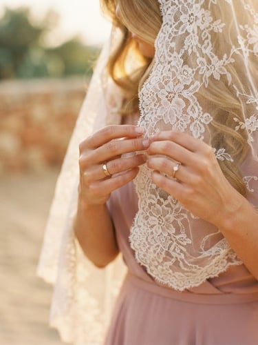 A close-up of a bride's hands adjusting a delicate lace veil, warm golden hour light filtering through, soft dusty rose tones, elegant editorial style.