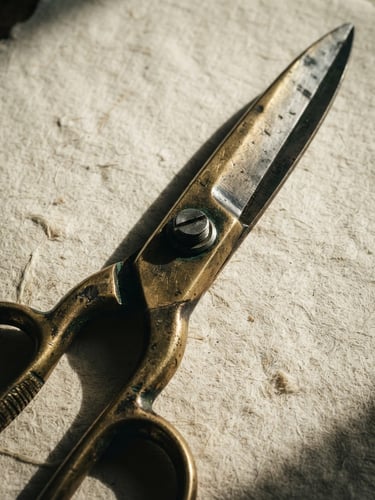 Extreme close-up of heavy brass shears resting on textured cream cotton paper, dramatic side-lighting, deep shadows
