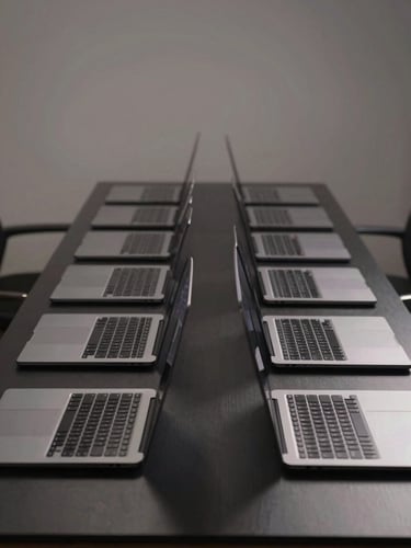 Symmetrical shot of a neat row of identical space gray MacBook Pros on a clean black workbench, edge-lit studio lighting.