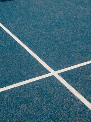 Close-up of a premium padel court surface at natural daylight, court lines sharp and precise against the blue-green playing surface, shallow depth revealing the texture and condition of the court, no players visible