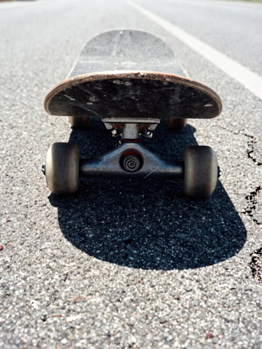 Low-angle close-up of scuffed skateboard wheels resting on a dusty, cracked asphalt highway shoulder, harsh midday sun glare, high-contrast desaturated tones.