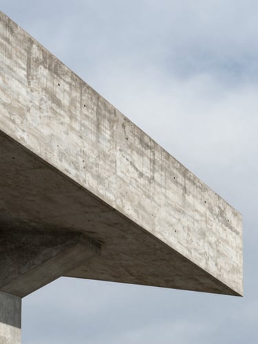 A sharp vertical close-up of a reinforced concrete cantilever beam projecting into empty space, cast shadows, overcast sky