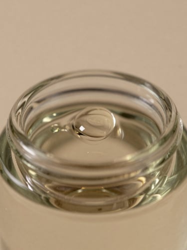 Macro close-up of a luxurious glass bottle reflecting soft studio light, rich oil droplet on the rim, warm neutral tones, high-end editorial, 85mm lens