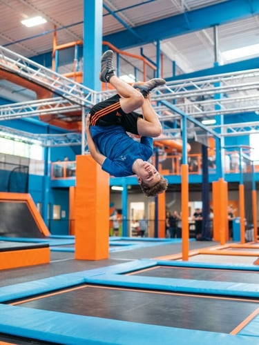 Dynamic low-angle action shot of a teenager mid-air performing a front flip on a trampoline, bright high-key indoor lighting, crisp 35mm, vibrant blue and orange park interior in background