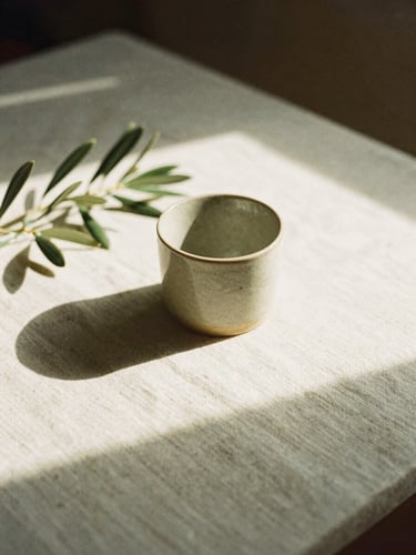 A sun-drenched minimalist workspace with soft shadows on textured linen paper, a ceramic cup, and delicate olive branches in morning light, 35mm film.