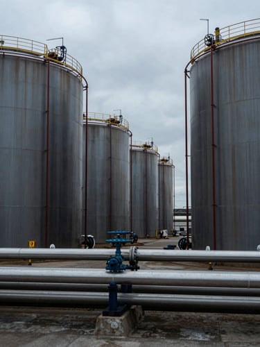Close environmental shot of a bulk liquid storage terminal at an industrial port, cylindrical tanks receding into the frame, overcast cool daylight, pipeline infrastructure in foreground, no people, muted steel and charcoal tones