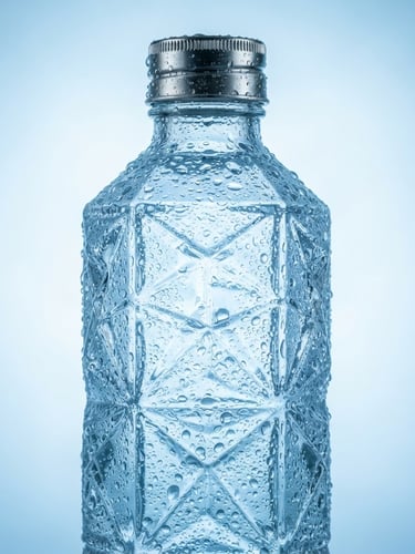 Studio photograph of a condensation-beaded premium glass water bottle, high-key cool blue backlighting, crisp focus on geometric glass facets, silver metallic cap.