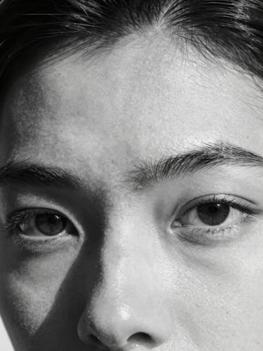 Stark black-and-white extreme close-up of a model's eyes and brow structure, high-contrast shadow cutting diagonally across the face, raw unretouched texture.
