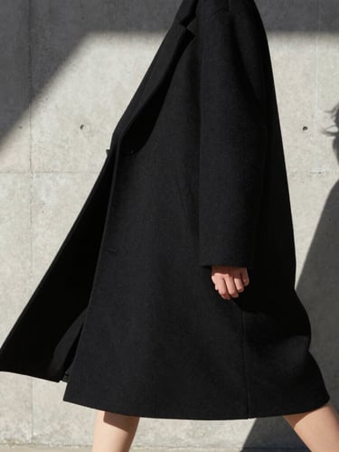 High-contrast architectural crop of a model in motion, wearing an oversized structural black coat, harsh direct sunlight, casting sharp geometric shadows on a concrete wall.