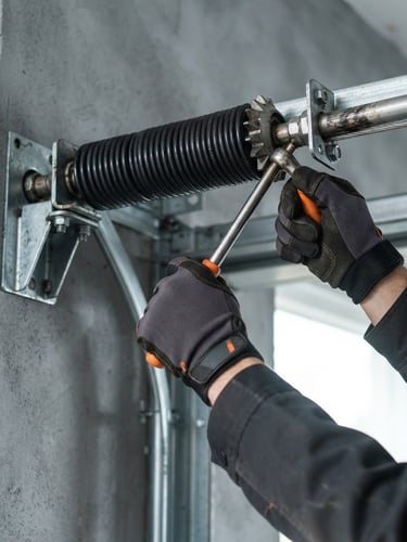 Close-up of a technician's hands in clean work gloves calibrating a heavy-duty torsion spring on a garage door, bright natural daylight, crisp industrial photography, safety-orange tools visible, slate gray background