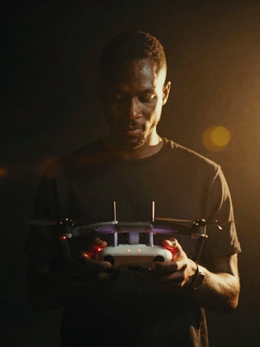 Low-key dramatic portrait of filmmaker Amalo Keita holding a drone controller, backlighting, deep shadows with subtle red record indicator light glow, cinematic