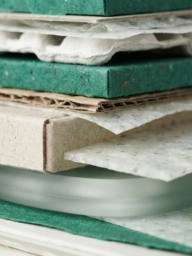 Extreme close-up of a stack of sustainable packaging materials, including recycled polymers, raw cardboard fibers, and glass, under bright studio lighting, sharp focus, clean emerald and white tones.