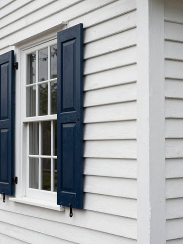 A close-up shot of a historic New England home siding being painted in crisp white, showing sharp paint lines against dark navy shutters under clear daylight.