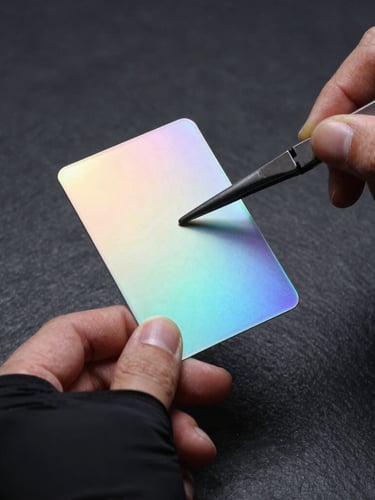Extreme macro studio photograph of hands in matte-black sleeves handling a pristine holographic card with tweezers under dramatic cyan side-lighting, dark slate background