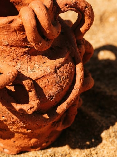 Extreme close-up of a vibrant terracotta sculptural vase under harsh direct sunlight, casting sharp dramatic shadows on a warm sand background, raw pigment textures visible, 35mm lens